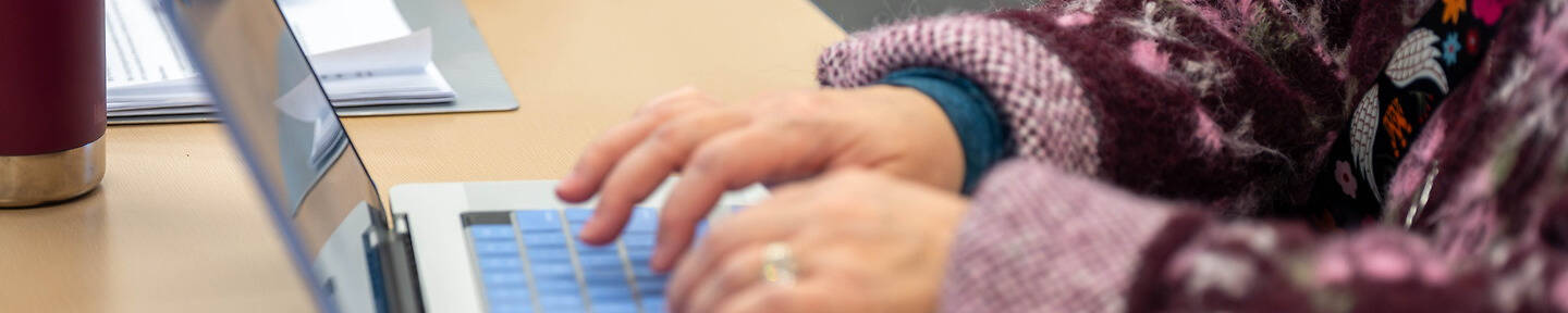 A person types on a laptop with a blue keyboard, wearing a patterned sweater, at a wooden desk beside a red travel mug and papers.